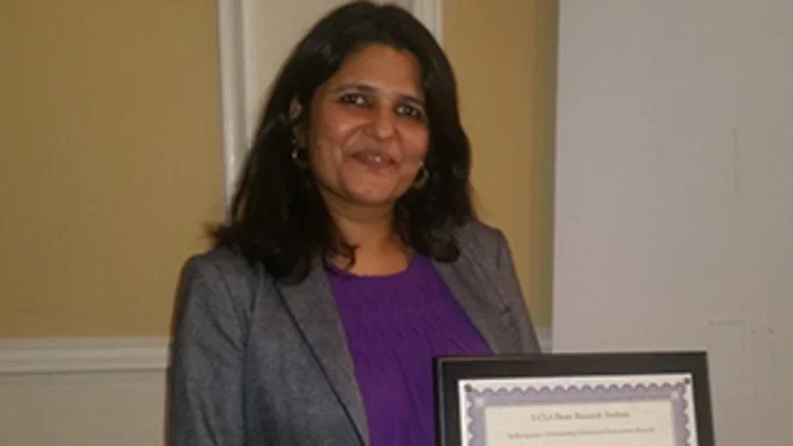 Woman in gray suit and purple shirt stands holding BRI post-doc award. 