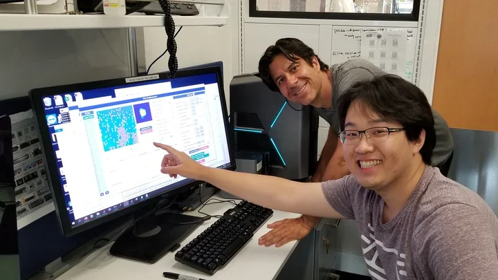 Two men smile at the camera while working at a computer in a lab.