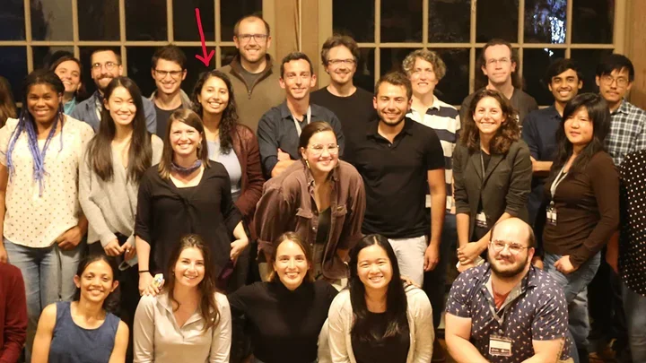 A group of people pose indoors for a group photo in front of large windows.