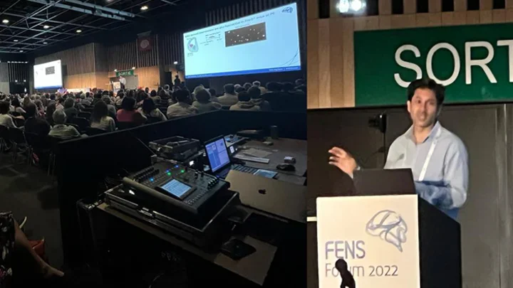 : A large audience sits in a darkened conference hall, facing a stage where speakers present slides on multiple projection screens.