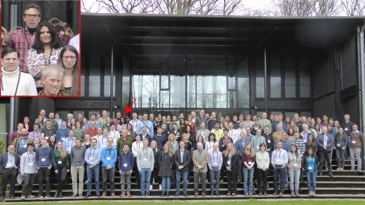 A large group of people pose for a group photo outside a modern building with large glass windows.