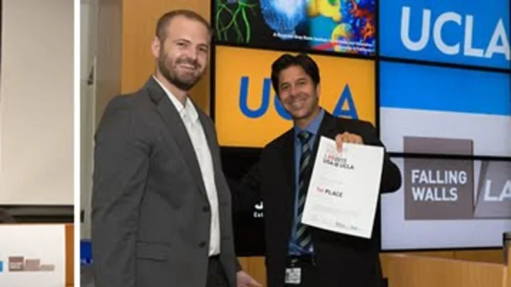 Two men in business attire stand in front of a screen displaying "UCLA." One man smiles at the camera while the other holds up a certificate and smiles.