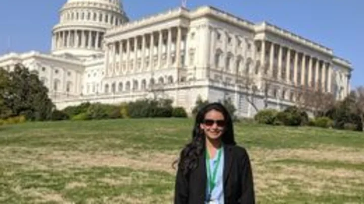A woman wearing sunglasses and a black suit stands smiling on the grass in front of the U.S. Capitol building on a clear, sunny day.
