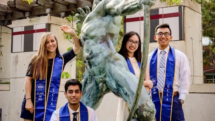 Four graduates wearing blue stoles pose around a bronze statue of Poseidon holding a trident.