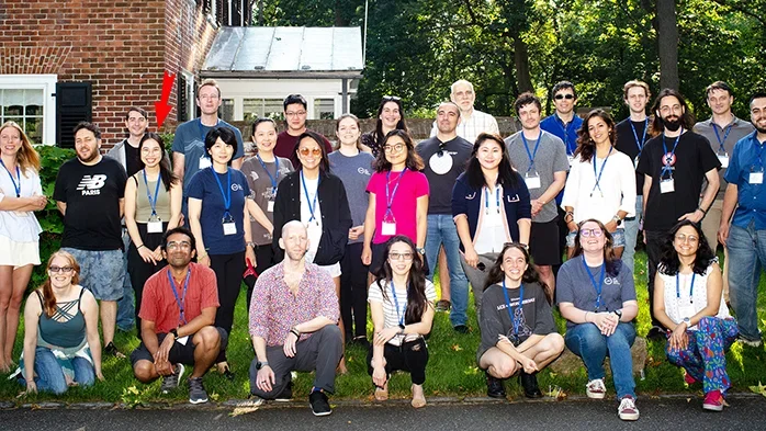 A group of people pose for a photo outside on a sunny day, standing and kneeling on grass in front of a brick building.