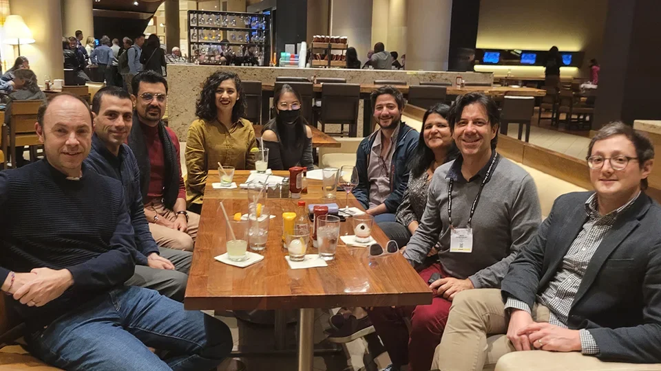 A group of people sit around a wooden table at a modern restaurant or hotel lounge, smiling for a group photo.