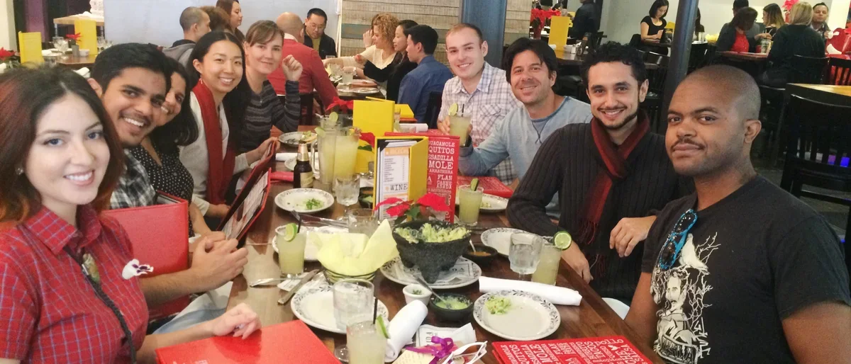 A group of people sit around a restaurant table, smiling and enjoying food and drinks.